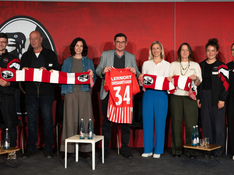 Menschen halten ein Trikot mit der Nr. 34 sowie einen Schal des SC Freiburg in die Kamera
